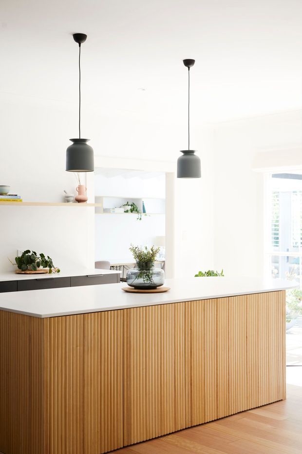 Bright contemporary kitchen centred on a large island with a white benchtop and vertically fluted timber cabinetry. Two dark pendant lights hang above the island, with open shelving, plants, and a sunlit windowed area in the background. The space has a clean, warm and minimalist feel.