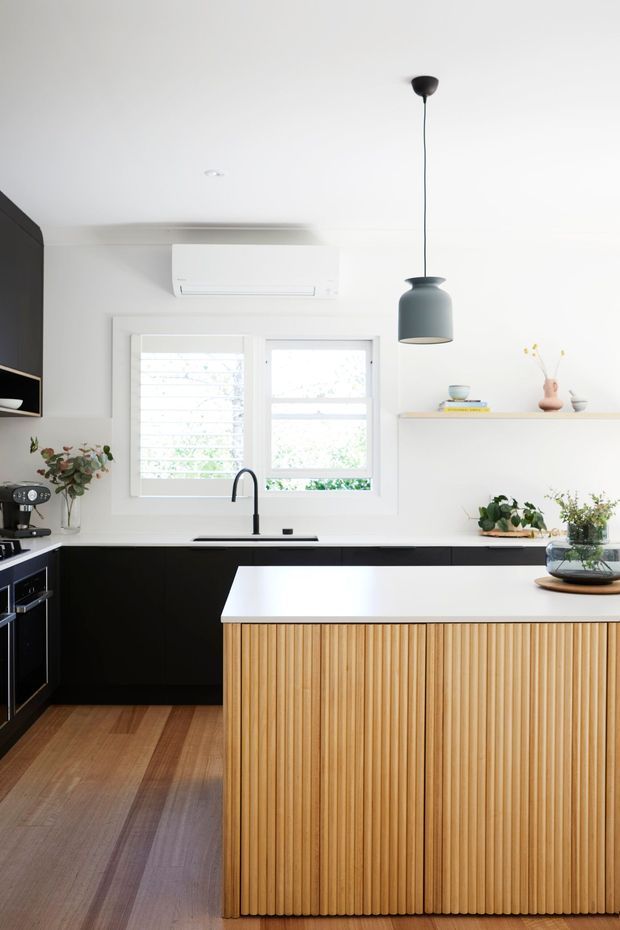 Bright contemporary kitchen featuring a large central island with a white benchtop and fluted light timber front, matte black cabinetry, timber flooring, white walls, a black gooseneck tap, and a dark pendant light. Plantation-shuttered windows and open shelving add natural light and understated styling.