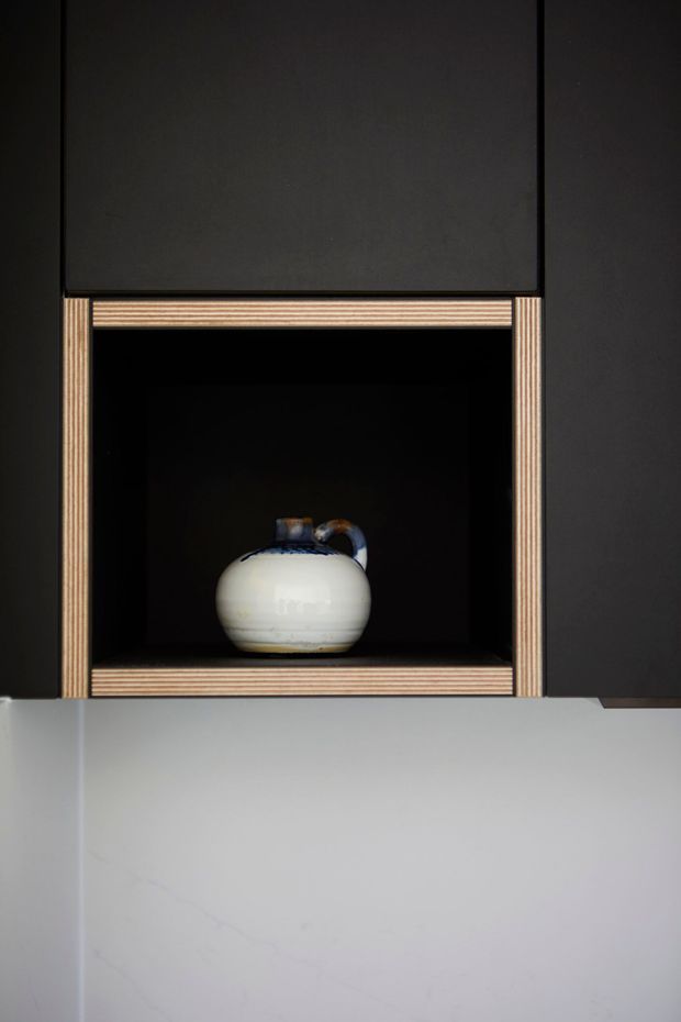 Close-up detail of a dark built-in kitchen or display joinery wall, featuring an open black niche framed with exposed layered timber plywood edges. A small ceramic vessel is displayed inside, above a pale stone or engineered-stone benchtop/splashback surface.