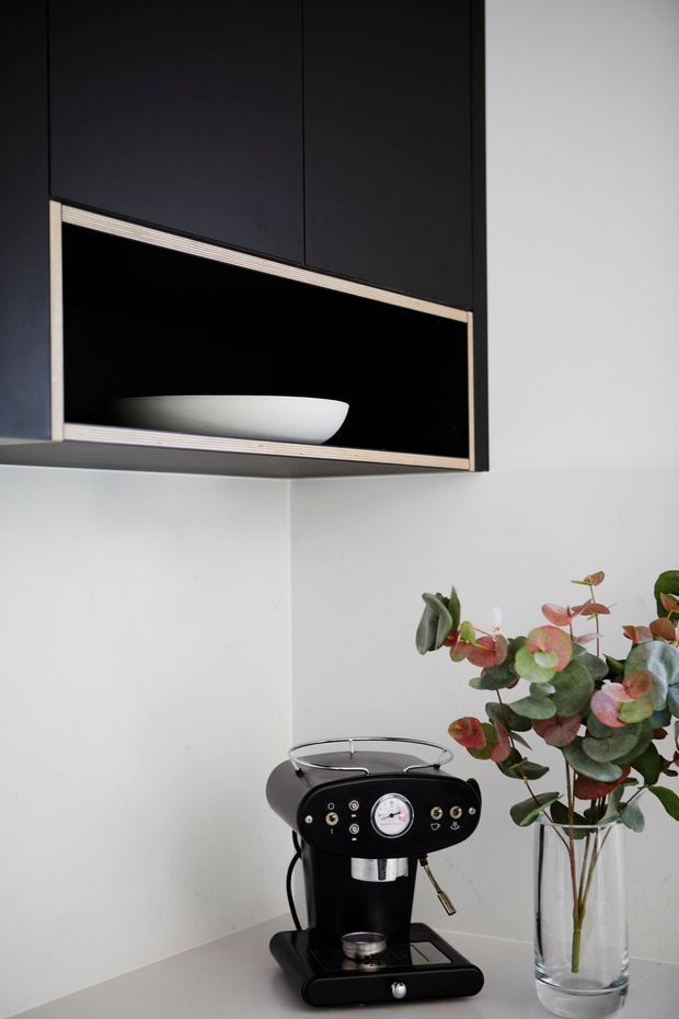 A minimalist kitchen coffee station with a black built-in overhead cabinet featuring an open display niche trimmed in light timber. A black espresso machine sits on a pale stone or composite benchtop beside a clear glass vase of eucalyptus foliage. The space has clean white walls, dark cabinetry, and restrained modern detailing.