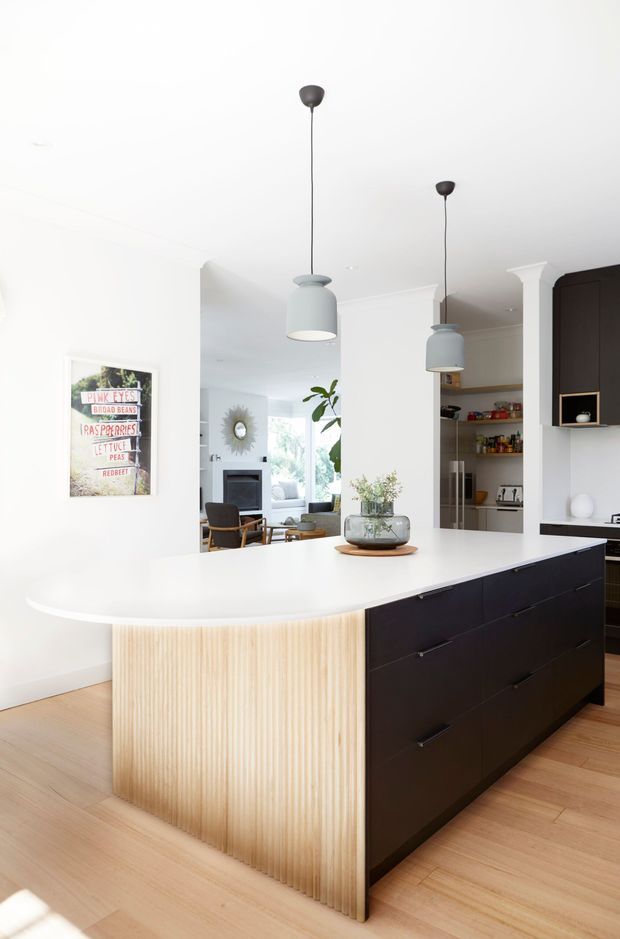 Bright contemporary kitchen featuring a large curved white island with a vertically fluted light timber end panel and dark cabinetry. Two pendant lights hang above the island, while a pantry, built-in cabinetry, and an adjoining living area are visible beyond. Light timber flooring, white walls, and black accents create a clean, refined interior.