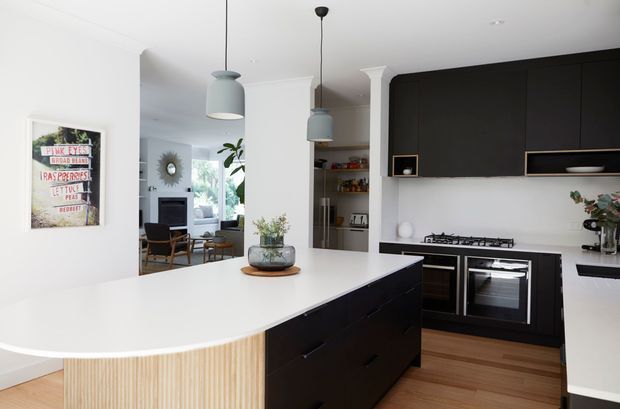 Contemporary open-plan kitchen with a large white waterfall island, dark matte cabinetry, integrated double ovens, gas cooktop, timber flooring, and pale blue pendant lights. The kitchen opens to a light-filled living area with a visible seating zone and decorative shelving.