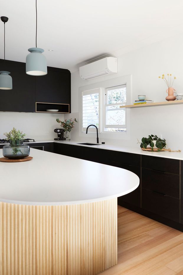 Contemporary kitchen featuring a large curved island with a white benchtop and vertically fluted light-wood front, paired with matte black cabinetry and integrated shelving. White walls and ceiling, timber flooring, black-framed fixtures, pendant lighting, a sink beneath a shuttered window, and restrained botanical styling create a clean, high-contrast interior.