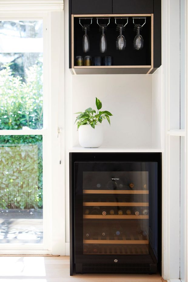A bright built-in kitchen or entertaining nook beside a tall window, featuring a black and brass-toned glassware cabinet with hanging wine glasses above an open white shelf, a potted plant, and a freestanding wine refrigerator below. White cabinetry and trim contrast with the dark appliances and light timber flooring.