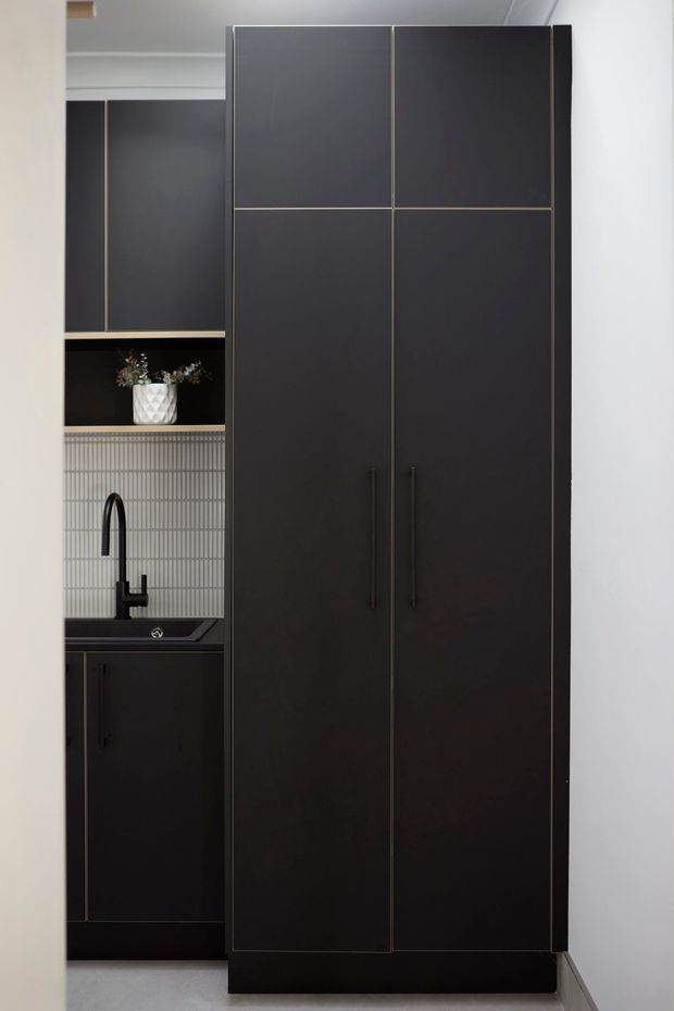 A compact, dark-toned kitchen or kitchenette viewed through a doorway. Floor-to-ceiling matte black cabinetry dominates the frame, with tall pantry-style doors, slim dark handles, a black sink and curved black tap, and a vertically ribbed light grey tiled splashback. An open shelf with a small vase provides a subtle decorative element. The space has a clean, refined contemporary feel with strong tonal contrast.