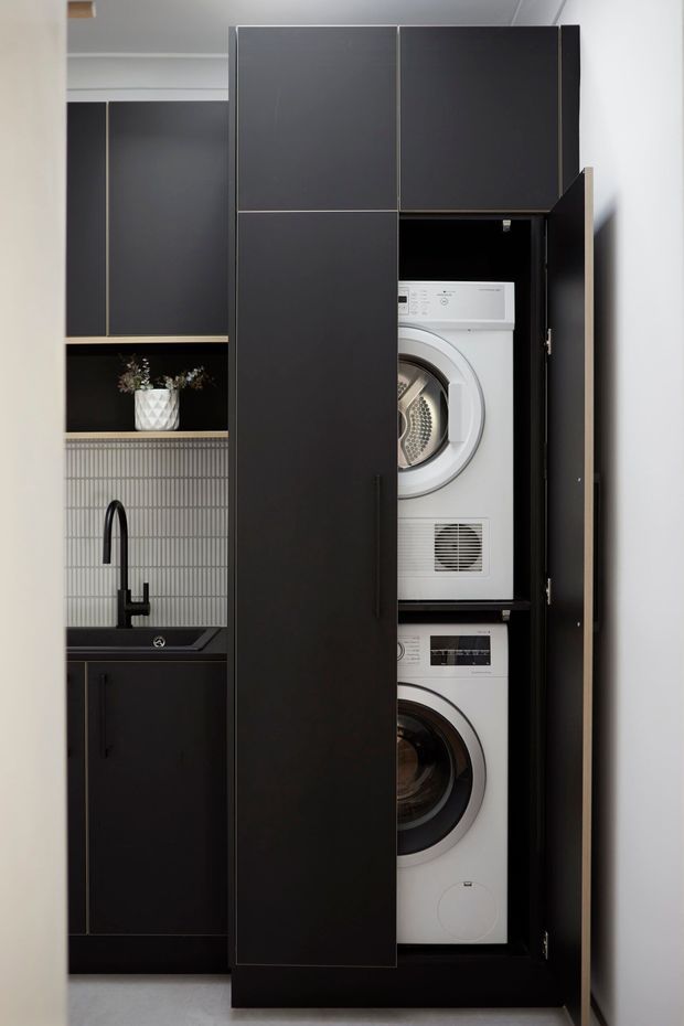 A compact, contemporary laundry room with a tall matte-black built-in cabinet concealing stacked white washer and dryer units. Beside it is a black sink and tap set into dark cabinetry, with a vertically ribbed grey tile splashback and open shelving displaying a small plant. The space has a clean, highly tailored joinery design and neutral palette.