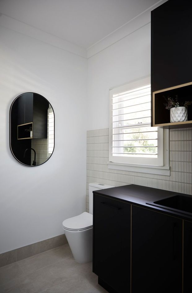 A compact contemporary bathroom with a white wall palette, pale grey floor tiles, vertically lined light wall tiles, a black vanity with fine brass detailing, integrated black basin, white toilet, arched black-framed mirror, and a louvred window providing natural light.