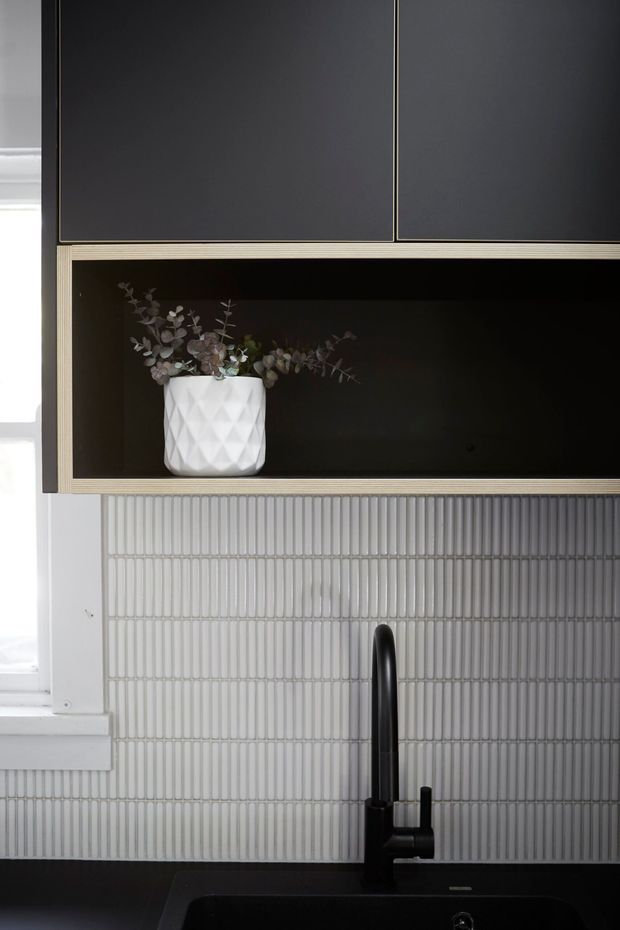 Close-up of a refined kitchen fit-out featuring dark matte overhead cabinetry, an open black display niche with a white faceted vase, vertically ribbed light-grey tiled splashback, and a matte-black gooseneck tap over a dark sink. A bright window is partially visible at the left.