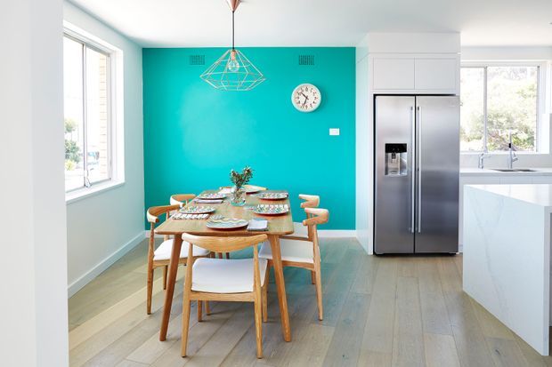 Bright open-plan dining area adjoining a modern kitchen. A light timber dining table with six matching chairs sits on pale timber flooring, backed by a vivid turquoise feature wall with a geometric pendant light and round wall clock. White cabinetry, a stainless-steel French-door refrigerator, pale stone island cabinetry, and large windows complete the space.