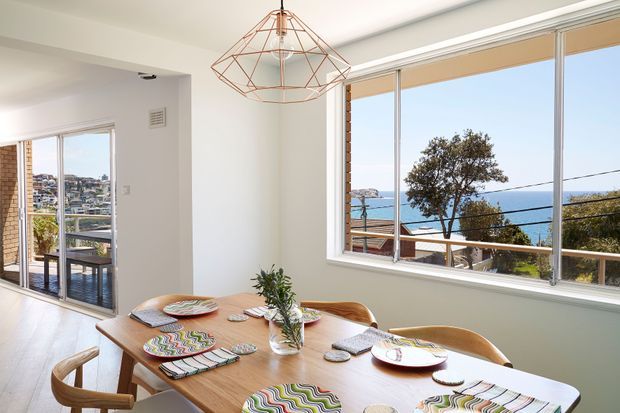 Bright dining area with a timber dining table, woven placemats, wooden chairs, and a geometric pendant light. White walls and large windows and glazed doors provide expansive views toward a blue coastal bay and surrounding hillside residences.