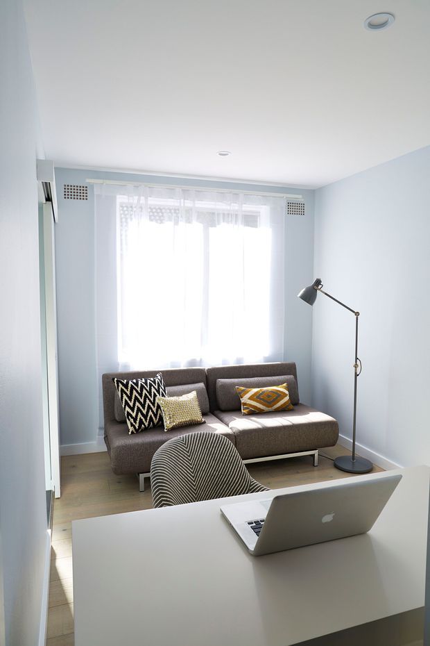A compact, bright multipurpose room arranged as a home office and sitting area. A white desk with an open laptop and patterned upholstered office chair occupies the foreground, while a brown two-seat sofa with geometric cushions sits beside a slim black floor lamp. Pale blue walls, white ceiling, sheer window curtains, light timber flooring, and recessed ceiling lights create an airy contemporary interior.