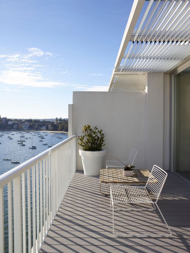 A bright contemporary waterfront apartment balcony or terrace with dark timber-look decking, white vertical balustrades, a white louvred pergola, and sliding glazed doors. Minimal outdoor furniture includes a slatted table and white wire-frame lounge chairs, with a potted plant beside the wall. A calm harbour with moored boats and a residential shoreline is visible beyond the balcony.