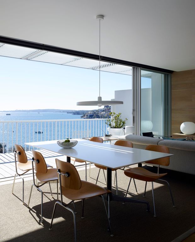 Bright, refined open-plan dining and living area overlooking a waterfront. A long white dining table with sculptural wooden chairs sits beside full-height sliding glazing and a balcony, with a grey sofa, pendant light, timber feature wall and indoor plant. The space has strong indoor-outdoor connection, abundant natural light and expansive water views.