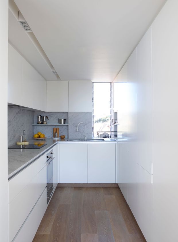 A bright, narrow contemporary galley kitchen with white flat-panel cabinetry, grey-white veined stone benchtops and splashback, integrated appliances, open shelving, stainless steel fixtures, and warm timber flooring. A tall window at the far end provides strong natural light and an urban outlook.