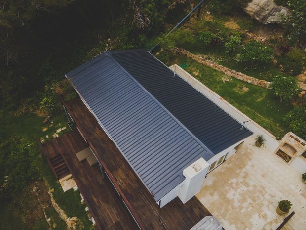 Aerial view of a contemporary single-storey building with a long dark ribbed metal roof, dark timber-clad exterior walls, and adjoining timber decking. A pale paved terrace and landscaped garden surround the structure.