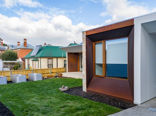 Exterior view of a contemporary single-storey extension beside a landscaped lawn and established neighbouring homes. The addition features a projecting box-like facade with a large timber-framed window, dark recessed reveal, rusty brown metal cladding, pale stonework, and smooth light-grey wall panels. A covered entry or connection leads toward the older dwelling, creating a clear contrast between modern and traditional forms.