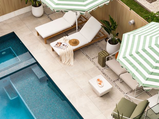 Elevated view of a stylish outdoor poolside terrace with a long rectangular swimming pool, pale tiled paving, white cushioned timber sun loungers, striped green-and-white umbrellas, potted tropical plants, and a warm vertical slatted privacy screen. The space feels polished, resort-like, and well suited to outdoor entertaining and relaxation.