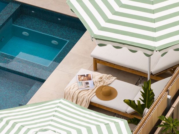 Elevated view of a stylish poolside terrace with a turquoise mosaic-tiled swimming pool, pale stone paving, striped green-and-white sun loungers, and a low outdoor daybed styled with a throw, hat, magazine, and tropical plants. The scene has a relaxed resort-like atmosphere.
