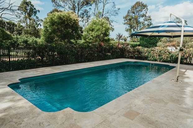 Outdoor swimming pool with clear blue water, surrounded by a pale stone-paved terrace, black metal fencing, lush garden planting, and a large striped sun umbrella. Mature trees form the background, creating a private landscaped setting.