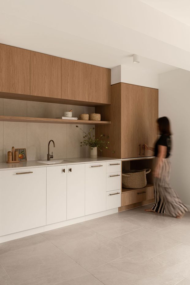 A warm, minimalist kitchen with a long run of white lower cabinetry, natural timber overhead cabinets and tall storage, a pale stone-look benchtop, and a matching tiled splashback. Open timber shelving displays ceramics and baskets, while integrated cabinetry and a woven basket add texture. The room has a light tiled floor, soft neutral tones, and simple ceiling lighting.