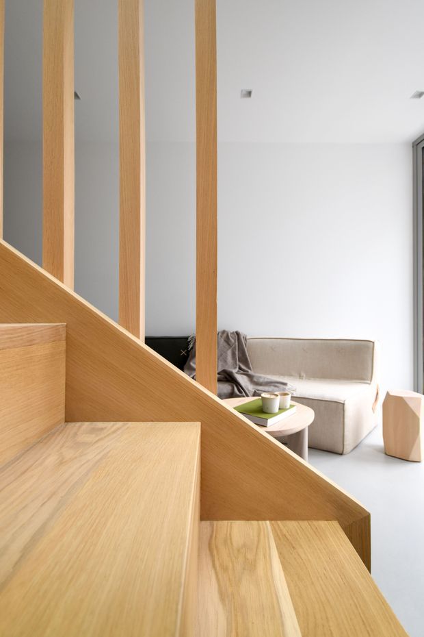 A bright, minimalist interior featuring a sculptural light-wood staircase with broad timber treads, a solid angled stair stringer, and vertical timber balusters. A compact neutral-toned sofa, round coffee table, and geometric timber side table are visible beyond the stair, with white walls, pale flooring, and recessed ceiling lights creating an airy contemporary setting.