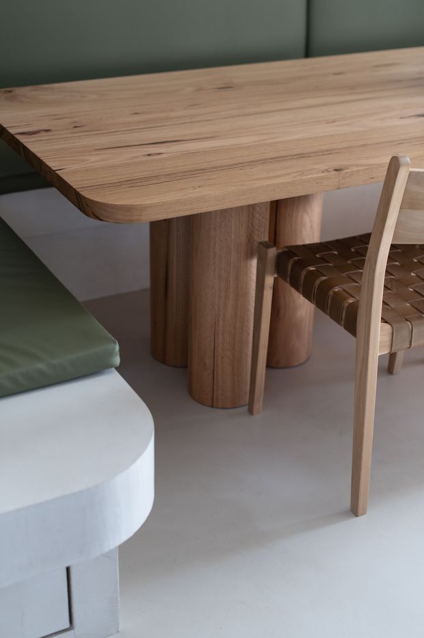 Close-up interior view of a light timber dining table with softly rounded corners and cylindrical timber legs, accompanied by a timber-framed chair with a dark woven leather or strap seat. A built-in upholstered bench with a muted green cushion and rounded white side table are partially visible. The scene has a restrained, contemporary Scandinavian feel with natural materials and soft neutral finishes.