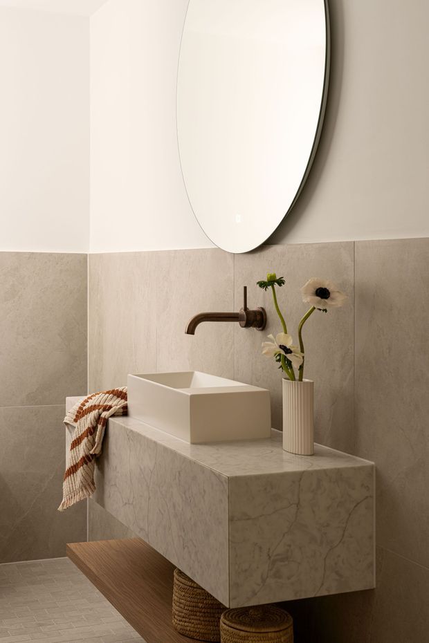 A refined contemporary bathroom vanity vignette with a floating marble-clad cabinet, square vessel basin, wall-mounted aged bronze tapware, large oval mirror, beige stone wall tiles, and a warm timber shelf with woven baskets beneath. A ribbed ceramic vase and simple flowers add a natural decorative touch.