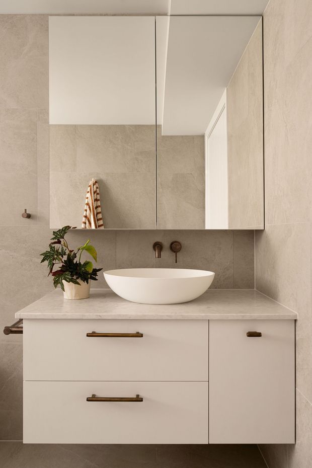 A refined contemporary bathroom vanity with a floating off-white cabinet, white vessel basin, pale stone benchtop, bronze tapware and handles, and large mirrored storage above. The walls and floor are finished in warm beige stone-look tiles, creating a calm, cohesive space.