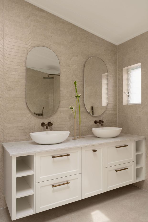 A refined double-vanity bathroom with a long white built-in vanity, two vessel basins, brushed bronze wall-mounted tapware, arched mirrors, and open shelving. The walls and floor are clad in warm beige textured tile, while a small frosted window provides natural light. The overall look is calm, warm, and contemporary with subtle classic detailing.
