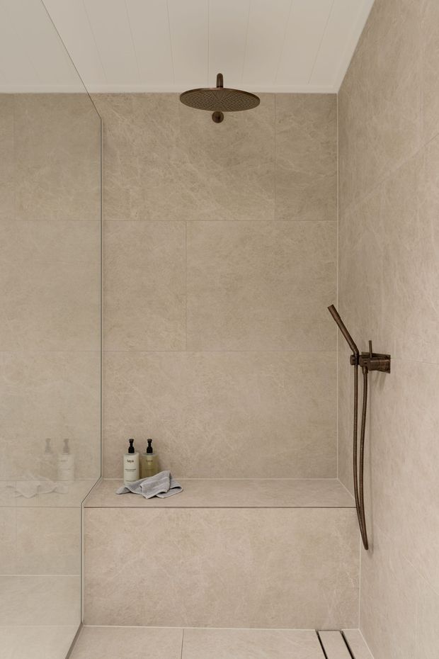 A minimalist walk-in shower clad in large-format warm beige stone-look tiles, with a built-in tiled bench, frameless glass screen, bronze rainfall showerhead and handheld shower, and recessed floor drainage. The white panelled ceiling and restrained fixtures create a calm, refined bathroom setting.