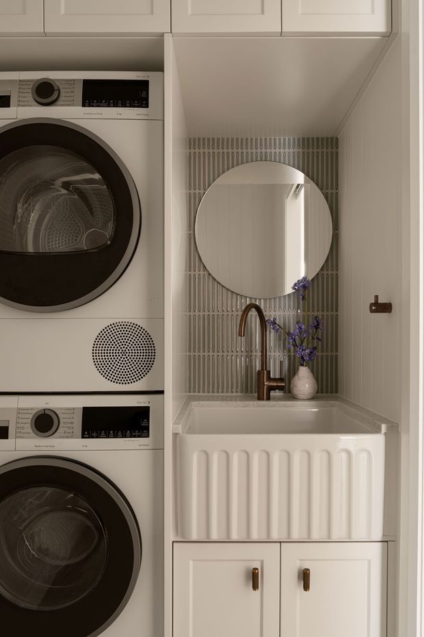 A compact, carefully fitted laundry room with stacked white front-loading washer and dryer on the left, and a white farmhouse-style utility sink with fluted apron front on the right. The sink area features vertically ridged wall tile, a round mirror, warm brass-toned tapware and hardware, white paneled cabinetry, and a small vase of purple flowers. The space has a refined, warm neutral palette and traditional detailing.
