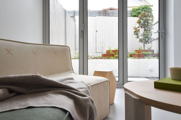 A softly lit contemporary living room viewed from beside a pale upholstered lounge chair, with a draped grey throw and muted green cushion. Full-height glazed doors open the room visually to a compact planted courtyard with raised garden beds and light vertical fencing. A rounded timber coffee table and small side table complete the calm, minimalist interior.