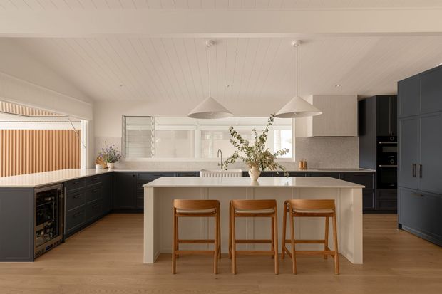 A bright, spacious contemporary kitchen with a large light-toned island and three timber bar stools. Dark navy cabinetry lines the perimeter, complemented by warm timber flooring, a pale benchtop, white vertically panelled ceiling, and two large white pendant lights. A built-in wine fridge is visible at the left, with open shelving and a window behind the sink. The kitchen has a warm, refined, clean aesthetic.