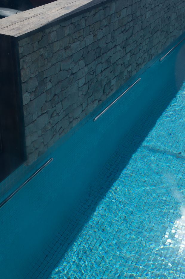 Close-up architectural photograph of a swimming pool beside a dark rough-stone masonry wall. The pool has blue mosaic tile lining, stainless-steel grab rails, and a timber-capped wall edge, with strong sunlight creating a high-contrast shadow across the water.