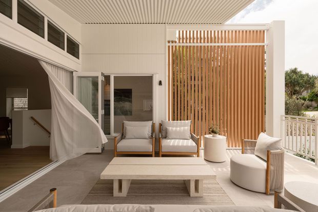 Bright contemporary covered outdoor lounge or sunroom opening directly to the garden, with white architectural finishes, a ribbed ceiling, timber screening, pale upholstered seating, a large coffee table, and a light neutral rug. Large sliding openings and curtains connect the space to the interior, while a white balustrade and landscaping are visible beyond.