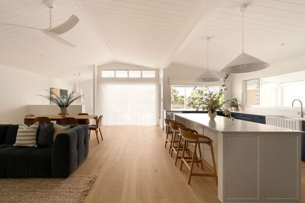 Bright open-plan coastal-style interior combining a living room, dining area and kitchen. The room features a white-painted vaulted timber-lined ceiling with exposed beams, light timber flooring, a large kitchen island with pale cabinetry and a white benchtop, dark navy lower cabinetry, pendant lights, ceiling fans, and generous glazing with sheer blinds. A dark upholstered sofa and timber dining furniture create a relaxed, warm residential setting.