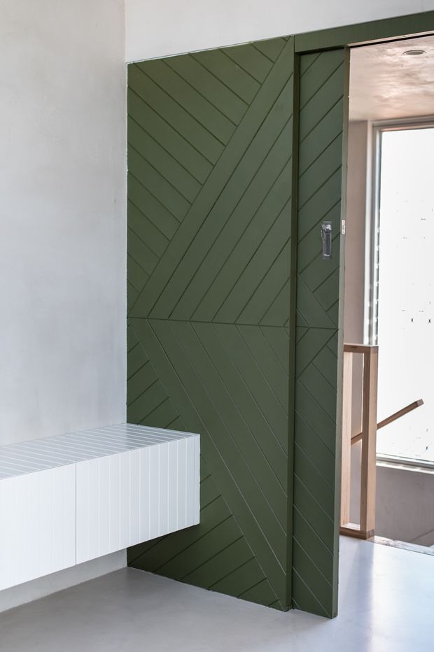 Interior view of a contemporary hallway or landing featuring a dark sage-green built-in wall and sliding door clad in diagonal chevron-patterned panels. A floating white built-in cabinet projects from the adjacent white wall, while an open doorway reveals a stair landing with timber handrails and bright glazing beyond. The space has a smooth grey concrete-like floor and a restrained, modern palette.