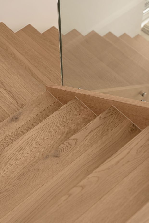 Close-up view of a contemporary timber staircase with broad oak-toned treads, matching timber risers and a solid timber side/guard detail. A clear glass balustrade reflects the staircase and adds a minimalist, open feel. The palette is warm and natural, with simple white walls and refined detailing.