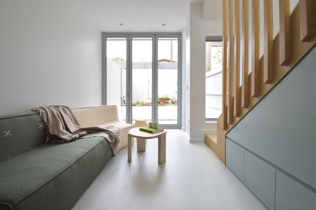 Bright, compact contemporary living room with a muted green sofa, small round timber coffee table, and a pale upholstered armchair. Full-height glazed doors open to a small enclosed outdoor area, while a timber staircase with integrated blue-grey cabinetry forms a strong architectural feature along the right side. The room has a clean, minimal palette and abundant natural light.