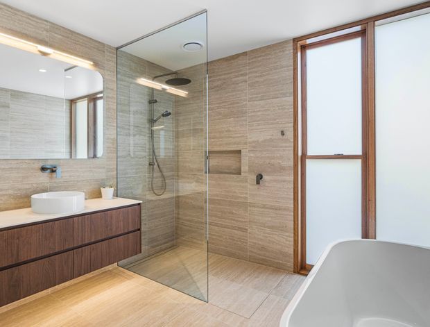 Contemporary bathroom featuring large-format warm beige stone-look wall and floor tiles, a frameless glass walk-in shower, floating dark timber vanity with a white vessel basin, wall mirror, and freestanding white bathtub. Timber-framed frosted glazing brings in soft natural light.