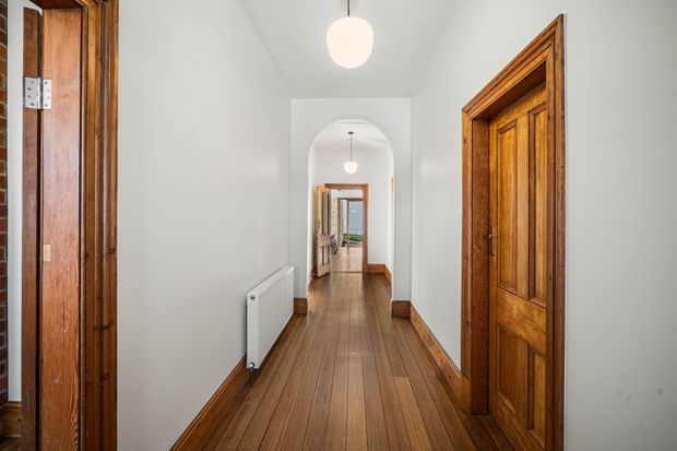 A long, narrow interior hallway with polished timber floorboards, timber skirting boards, and multiple warm-toned timber panelled doors with substantial trims. White walls and ceilings create a bright, clean corridor, with arched openings and simple pendant lights leading toward rooms at the far end.