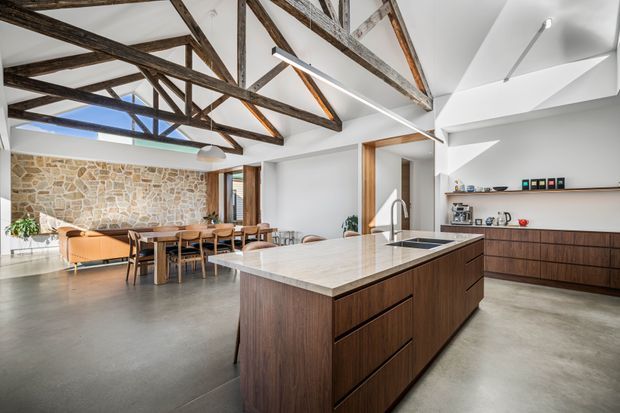 Open-plan kitchen and dining area with a large dark timber island, pale stone benchtop, integrated cooktop and sink, polished concrete flooring, and open shelving. The room features exposed rustic timber roof trusses, a rough natural stone feature wall, white angular ceiling forms, timber-framed openings, and abundant natural light.