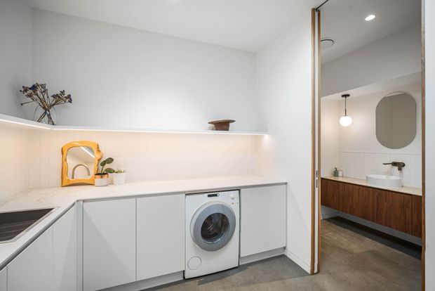 A bright, minimalist utility room with a compact white L-shaped kitchenette and integrated front-loading washing machine. Pale cabinetry, a light tiled backsplash, slim under-shelf lighting, and a small sink are visible. Through the open doorway is a contemporary bathroom with a floating dark timber vanity, white basin, rounded mirror, and pendant light. Grey flooring and white walls create a clean, restrained palette.