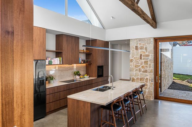 Bright, spacious contemporary kitchen with a large stone island and integrated sink, dark timber cabinetry, stainless-steel refrigerator, and built-in appliances. A suspended linear pendant hangs above the island. The room features a high white ceiling with exposed heavy timber beams, clerestory glazing, a natural stone feature wall, and large timber-framed glazed doors opening to an exterior courtyard. Bar seating accommodates several people.