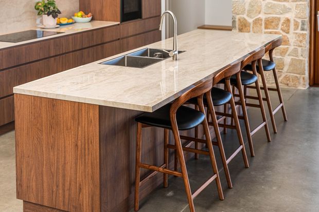 A contemporary kitchen dominated by a large island with a pale veined stone benchtop, integrated double sink, and brushed metal gooseneck tap. The island has dark timber-look cabinetry and is paired with several timber bar stools with dark seats. Matching dark timber cabinetry runs along the rear wall, with a black induction cooktop, while a rustic light stone feature wall adds texture. The space has polished concrete flooring and a warm, refined material palette.