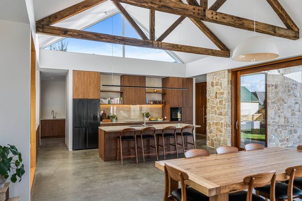 A bright open-plan kitchen and dining area with a large island, dark timber cabinetry, integrated shelving, black appliances, and warm wood furnishings. The room features a high vaulted ceiling with exposed rustic timber trusses, a large skylight or clerestory glazing, polished concrete flooring, and a prominent natural stone feature wall around the glazed doors. A timber dining table occupies the foreground, with views toward an exterior courtyard or neighbouring building.