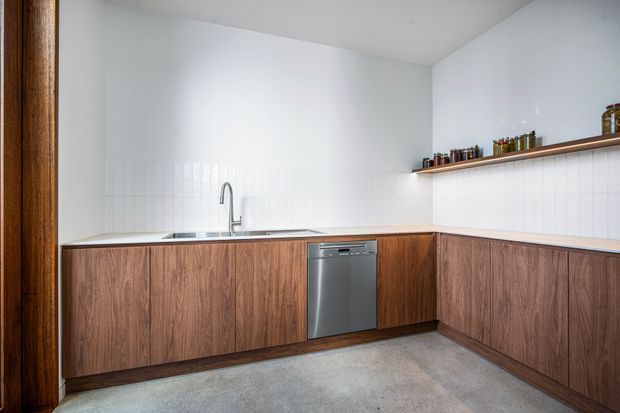 Bright, minimalist L-shaped kitchen with warm wood-grain base cabinetry, a stainless-steel dishwasher, white vertically tiled splashback and walls, a pale concrete-look floor, and a long open timber shelf displaying jars. A timber-framed opening is visible at left.