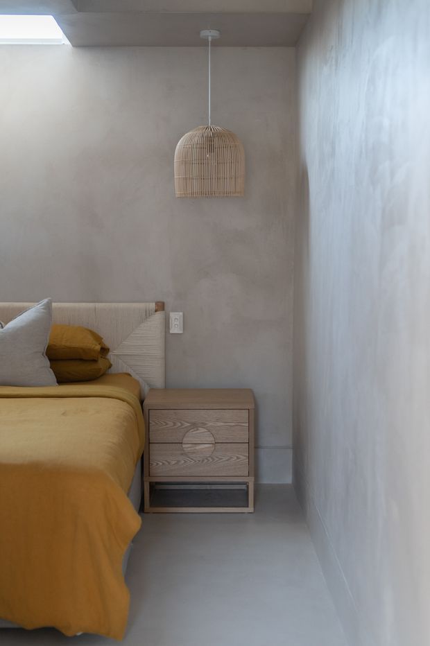 A compact, softly lit bedroom corner with a mustard-gold bedspread and pillows, a textured upholstered headboard, a light timber bedside table, and a woven pendant light. The walls and floor have a muted grey-beige plastered finish, with a skylight bringing in natural light. The space has a calm, warm, minimalist aesthetic.