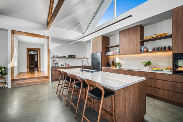 Bright open-plan kitchen dominated by a large stone island with seating for four, dark timber cabinetry, integrated appliances, open shelving, and a pale textured backsplash. The room has polished concrete flooring, a high sloped ceiling with exposed timber beams, clerestory glazing, and a connecting entry hall with timber-framed doorway visible in the background.