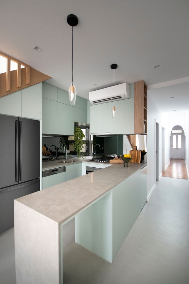 A bright contemporary kitchen with a long peninsula island, pale sage-green flat-panel cabinetry, and a light stone benchtop. The kitchen includes a large dark refrigerator, integrated dishwasher, gas cooktop, stainless sink, mirrored splashback, timber open shelving, pendant lights, and a wall-mounted air conditioner. A light hallway with timber flooring and an arched glazed door extends beyond the kitchen.